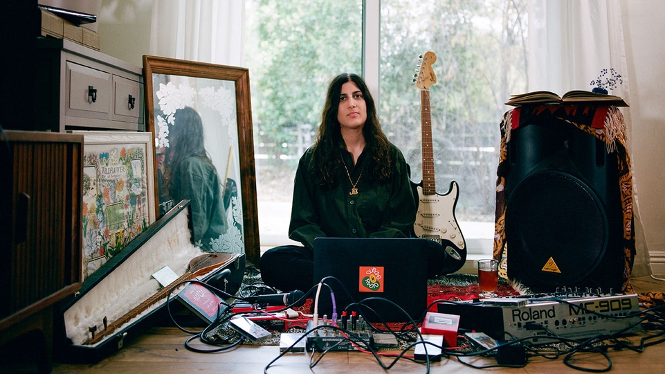 Maral sitting in front of a glass door, her fender stratocaster at her back and her studio equipment laid out on the floor in front of her