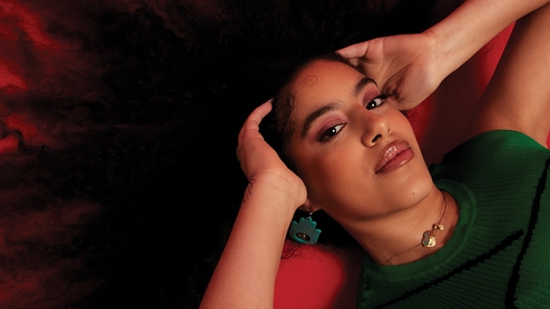 Press shot of Nooriyah lying on a red floor. Her curly hair is sprawled across the floor and she is holding either side of her head. She's wearing a green sleeveless top and green earrings