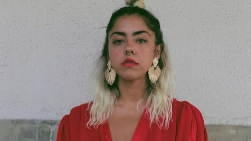 Photo of Paurro sitting against a white wall in a red robe. She has red lipstick on and ornamental earrings