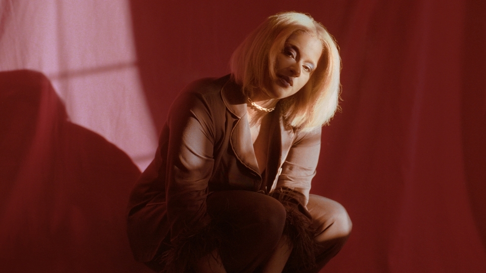 Photo of L’Rain crouching in a red dress in front of a shadowy red background