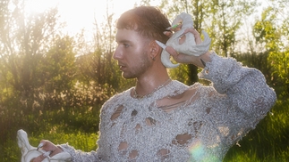 Photo of Bakblivv standing in a clearing of trees, the sun shining through the branches. He's wearing a ripped white top and has long acrylic nails