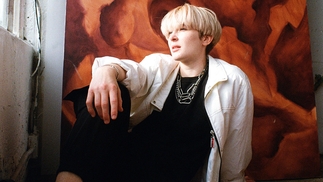 Ani Klang sitting on the floor with her arm resting on her knee. She's in front of an orange and red hanging abstract backdrop and is looking out the window