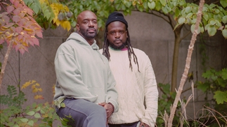 Both members of Black Cadmium standing side by side against a wall covered in hedge leaves