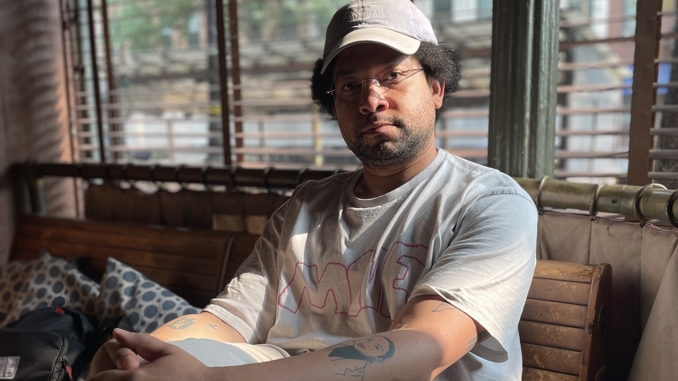 Photo of DJ Autopay sitting on a sofa while wearing a grey t-shirt and cream baseball cap