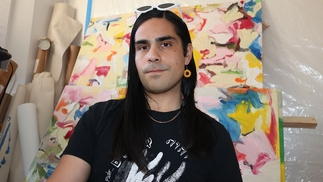 Photo of Farsight sitting in front of a colourful abstract painting. His hair is long and black and he has sunglasses resting on his head. He's wearing a dangling orange earing. 