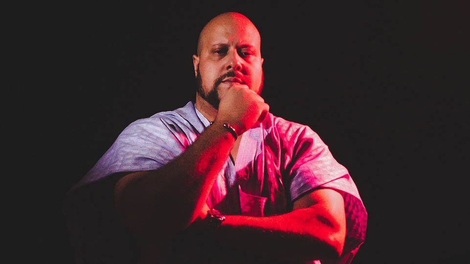 Boddhi Satva wearing a robe, photographed with his arms folded on a dark backdrop under a purple light 