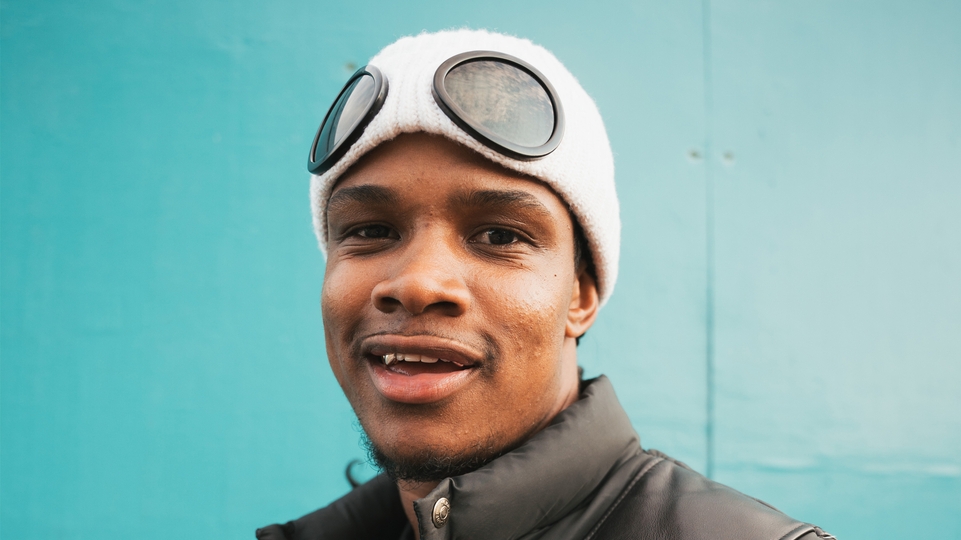 Photo of Kam-Bu posing wearing a white hat in front of a turquoise background