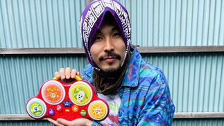 Photo of KASAI crouching in front of a light blue metal fence. He's wearing a patterend blue shirt and has a headscarf on. He's holding a child's toy musical instrument