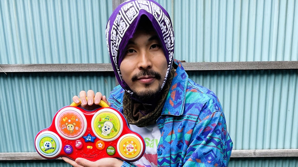 Photo of KASAI crouching in front of a light blue metal fence. He's wearing a patterend blue shirt and has a headscarf on. He's holding a child's toy musical instrument