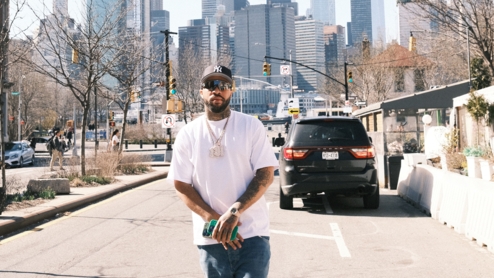 Larry June standing in front of a city skyline, wearing a hat, mirrored sunglasses, a white t shirt and blue jeans. He's holding two phones in his hands, and a black SUV is parked next to him