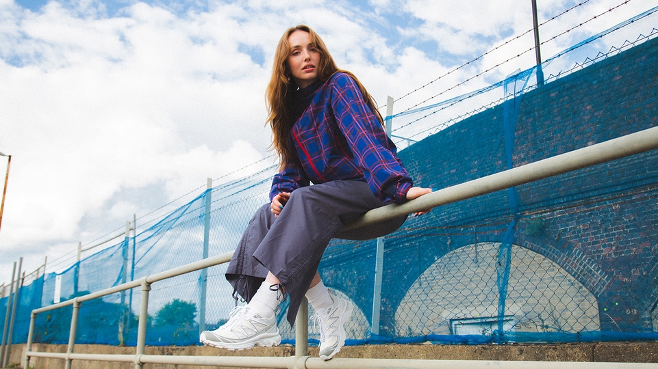 LUXE sitting on a steel bar fence in front of a blue mesh fence near some railway arches. She's wearing a dark blue top with red horizal and vertical stripes, grey blue trousers and white trainers