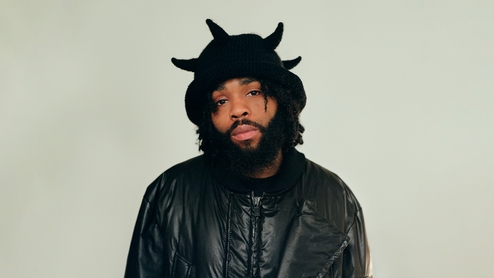 Fly Anakin wearing a black woolly hat with spikes on and a black coat. He looks straight into the camera and is stood in front of a grey background.