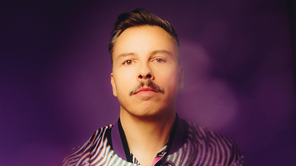 Purple Disco Machine looking into the camera against a purple background, surrounded by smoke