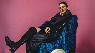 Saoirse reclining on a stool in a long blue velvet coat and knee high black boot