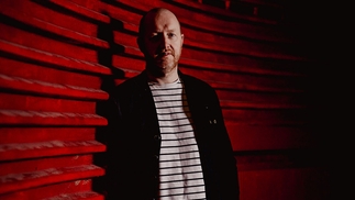 Phil Kieran in a black and white stripy t-short and cardigan leaning against a red surface