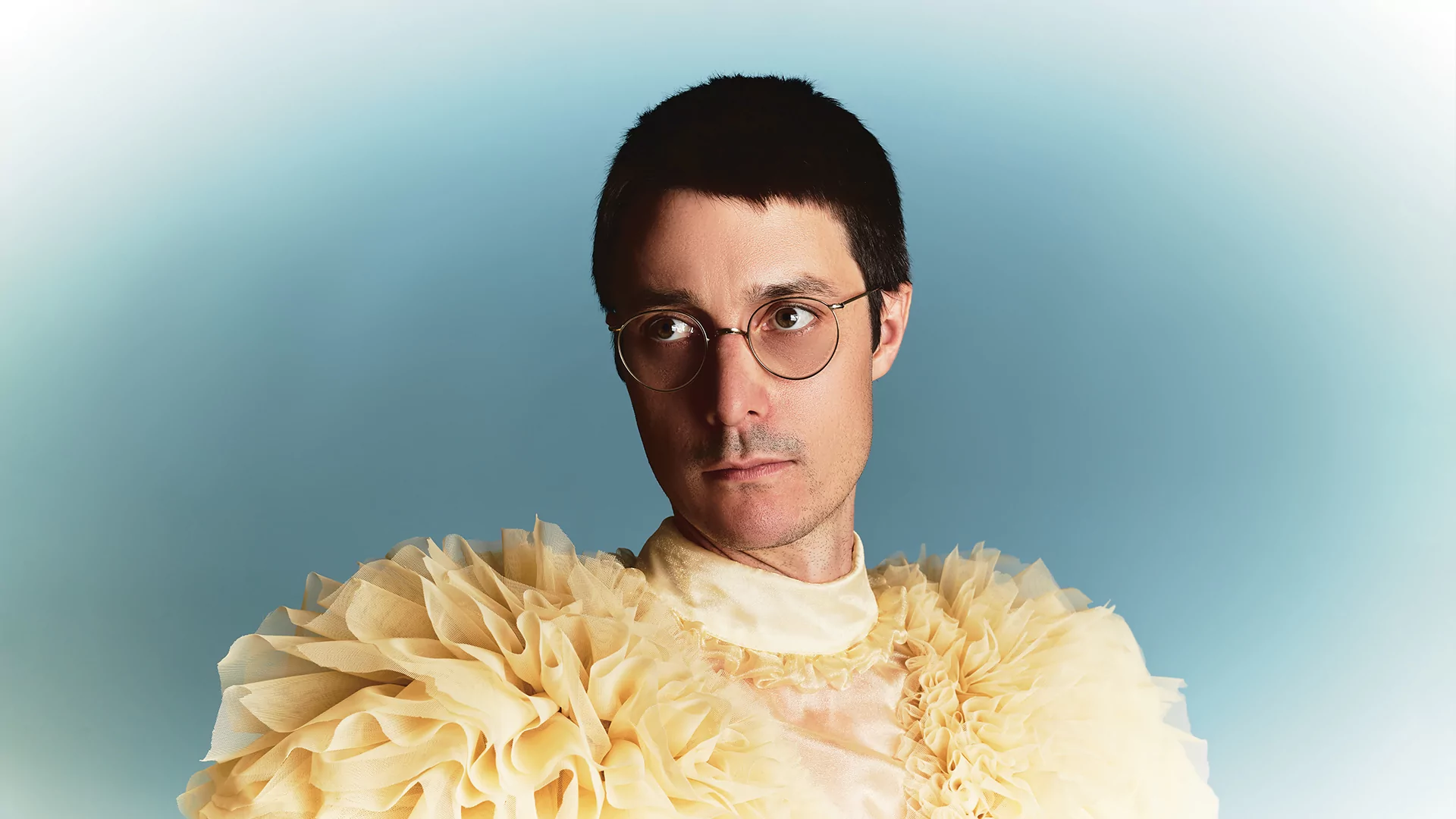 Call Super posing in glasses and a yellow ruffed top against a blue background