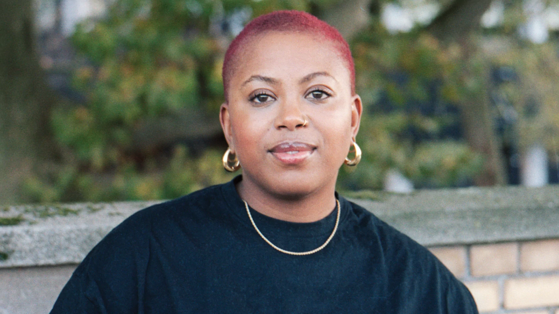 Press shot of Deborah X standing in front of a brick wall and a hedge. Her short hair is dyed red. 