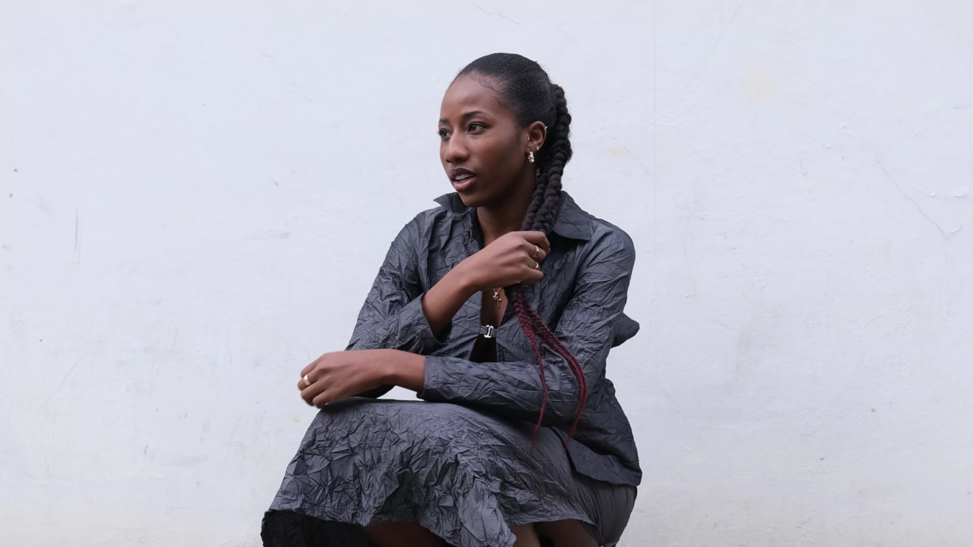 LORA crouching in front of a grey wall in a long dark grey coat