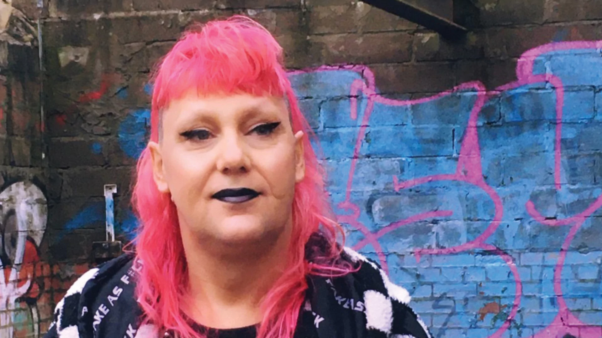 Gynoid 74 with long pink hair and black lipstick, standing in front of a brick wall covered in blue and pink graffiti