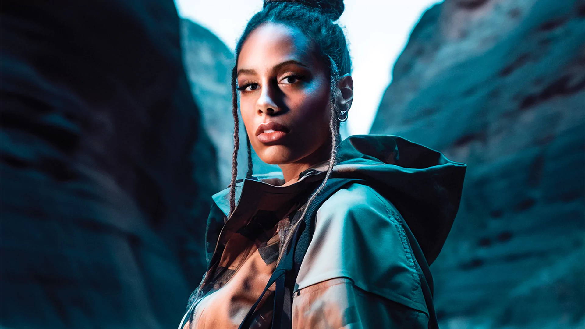 AI-edited photo of Jayda G looking over her left shoulder to the camera. She's wearing a dark puffer jacket and is between two ridges of a blue-lit valley