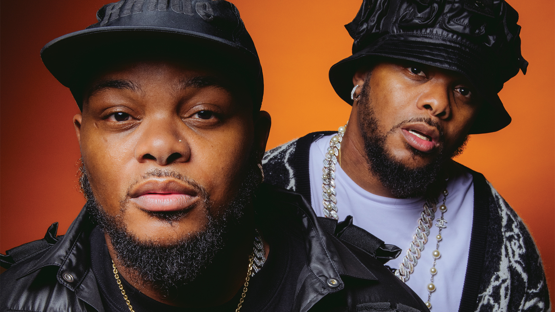 Photo of Bandile and Banele posing wearing black hats in front of a bright orange background