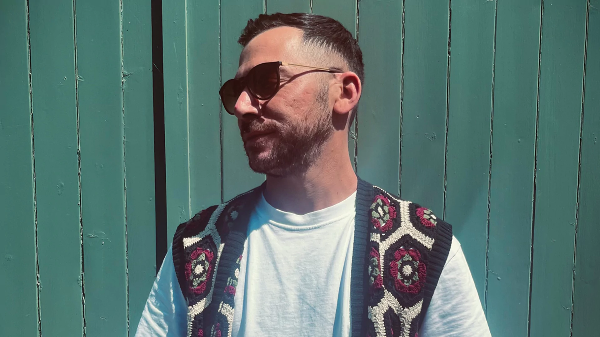 Max Sinàl posing in front of green wall. He's looking to the left of the camera and wearing shades