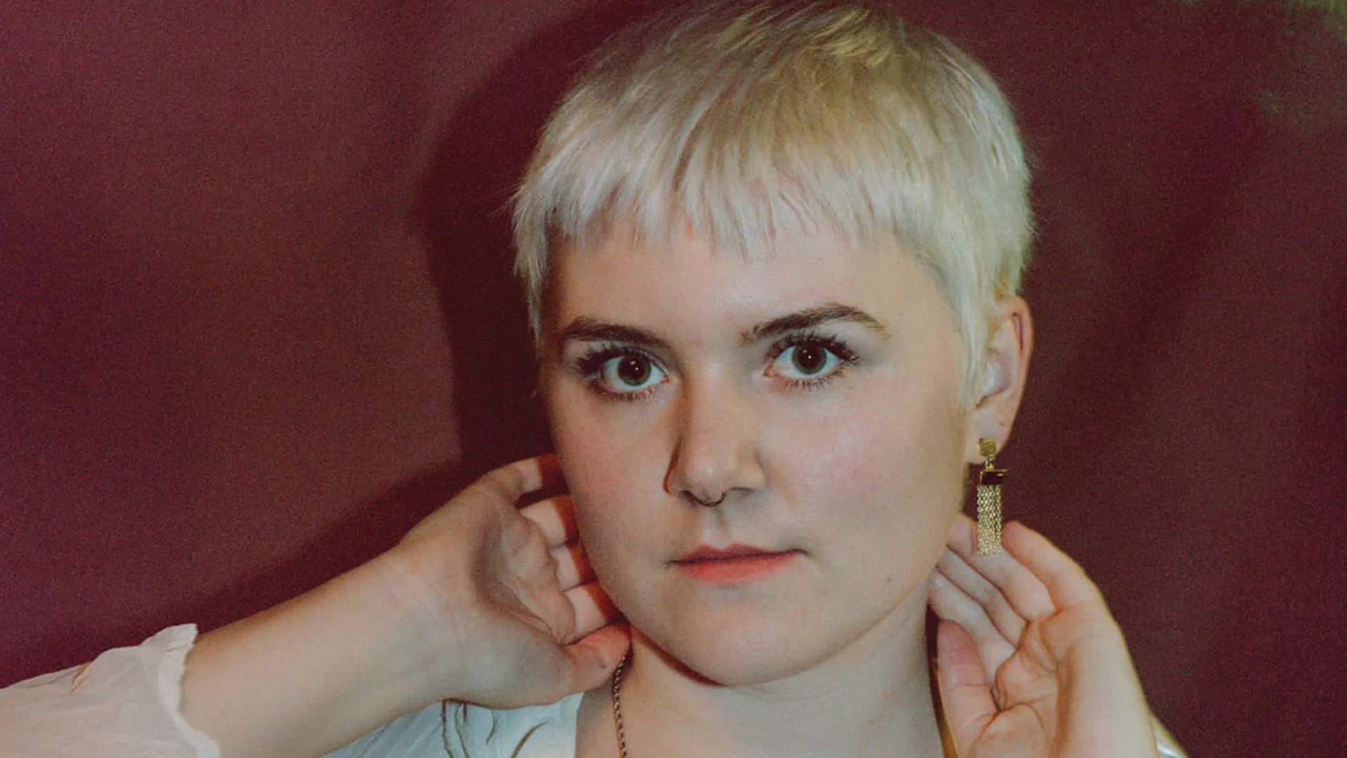 Percorn posing in front of a faded red curtain. Her hands are reaching behind her neck. Her hair is short and bleached blonde