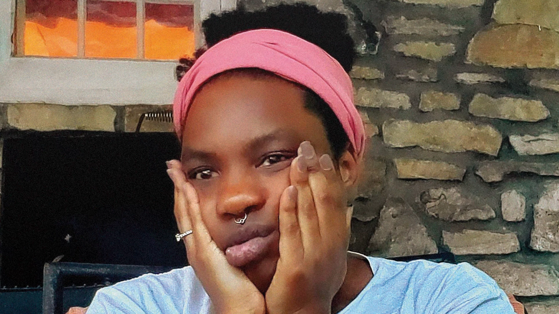 Photo of Nondi_ in front of a brick wall and fireplace. Her hair is held back with a pink headband and her face is resting in her hands